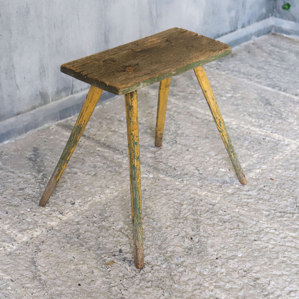 A small antique rustic timber milking stool featuring four angled splayed legs and a distressed yellow and green painted finish. View VIII