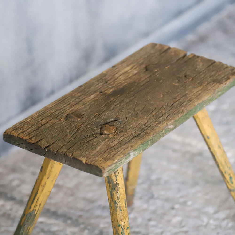 A small antique rustic timber milking stool featuring four angled splayed legs and a distressed yellow and green painted finish. View IX