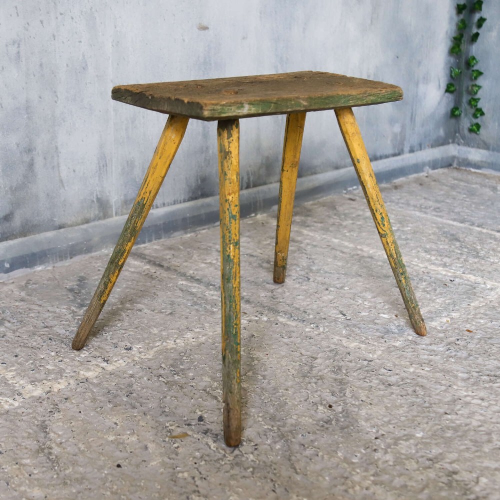 A small antique rustic timber milking stool featuring four angled splayed legs and a distressed yellow and green painted finish. View X