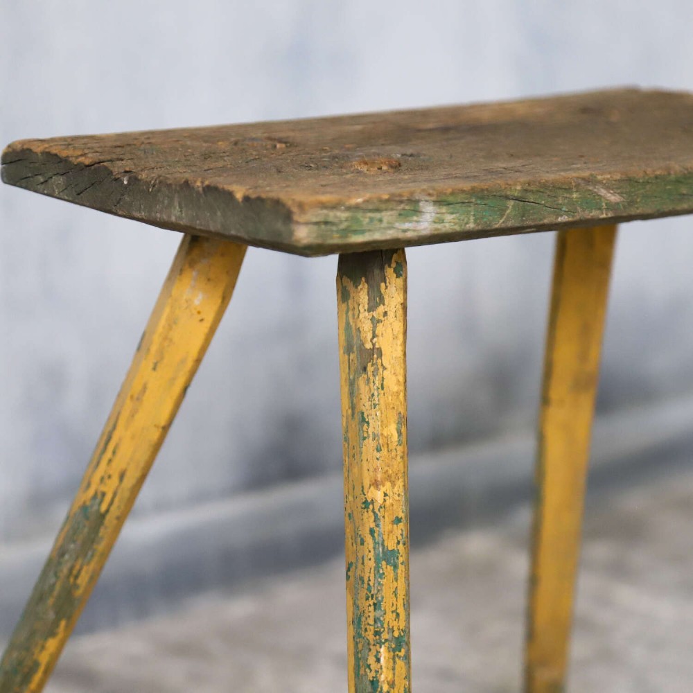 A small antique rustic timber milking stool featuring four angled splayed legs and a distressed yellow and green painted finish. View XI