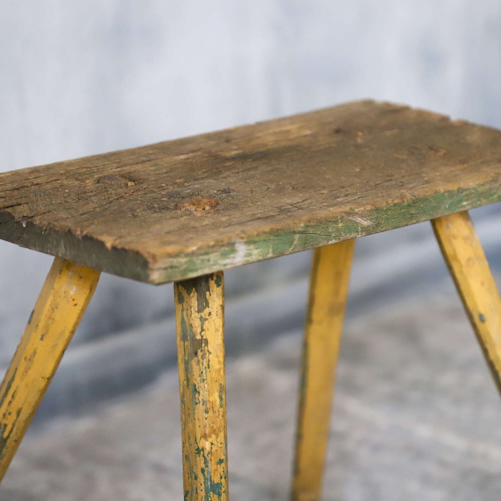 A small antique rustic timber milking stool featuring four angled splayed legs and a distressed yellow and green painted finish. View XII