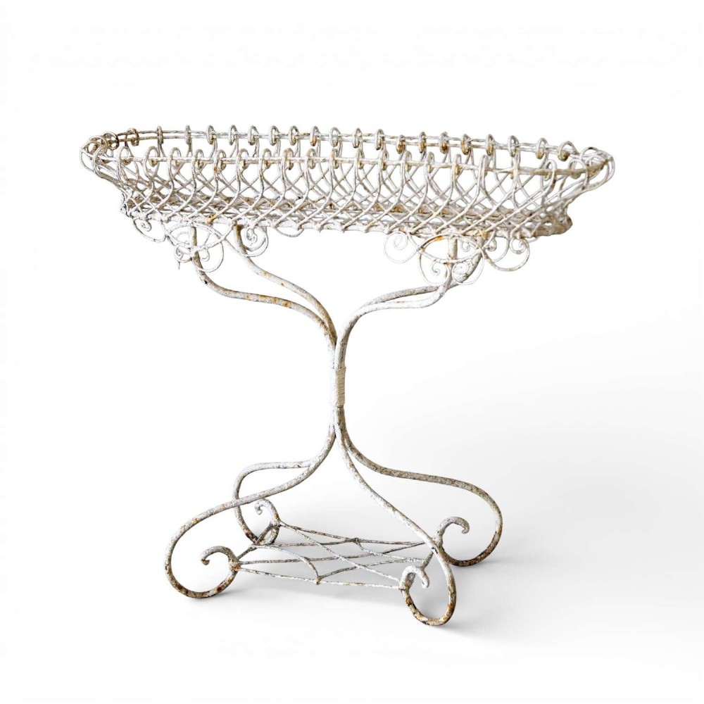 A 19th-century distressed white painted wrought iron plant stand featuring a wire lattice basket top over scrollwork legs.