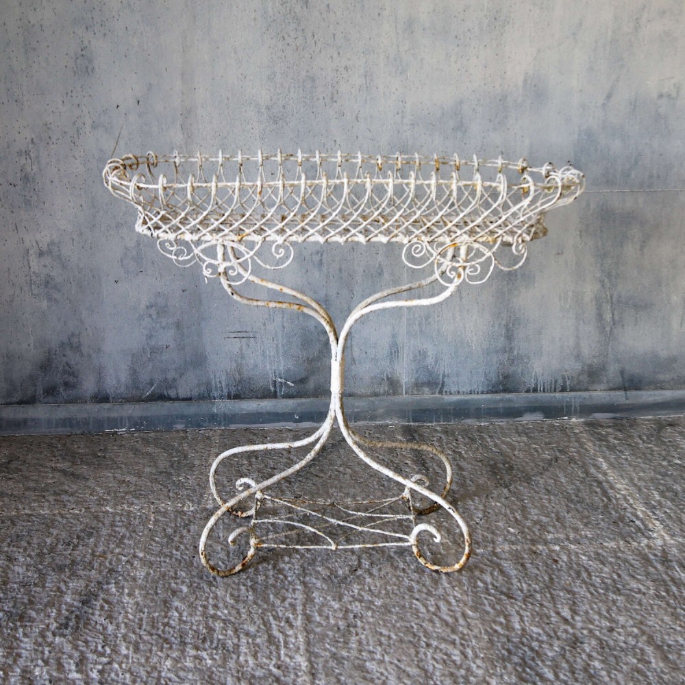 A 19th-century distressed white painted wrought iron plant stand featuring a wire lattice basket top over scrollwork legs. View II
