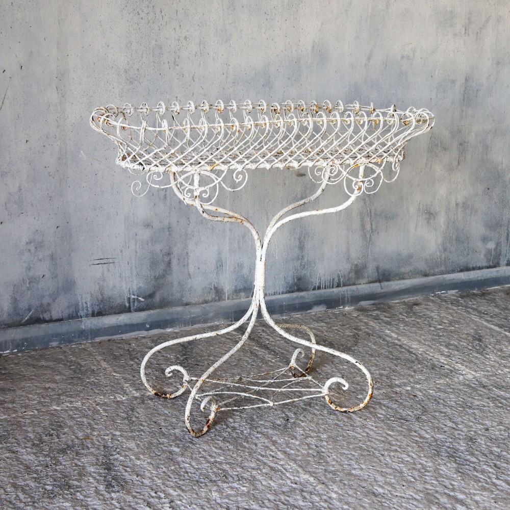 A 19th-century distressed white painted wrought iron plant stand featuring a wire lattice basket top over scrollwork legs. View III