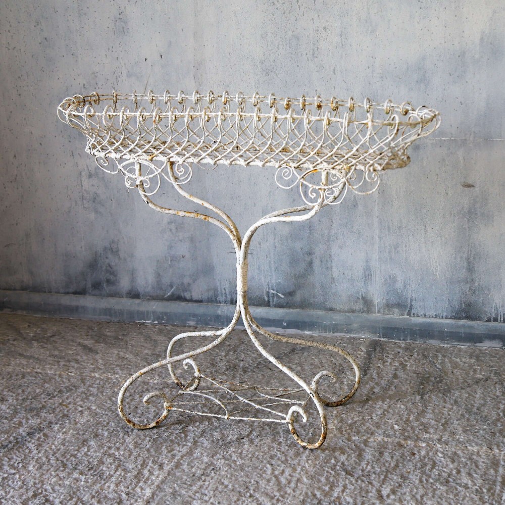 A 19th-century distressed white painted wrought iron plant stand featuring a wire lattice basket top over scrollwork legs. View IV