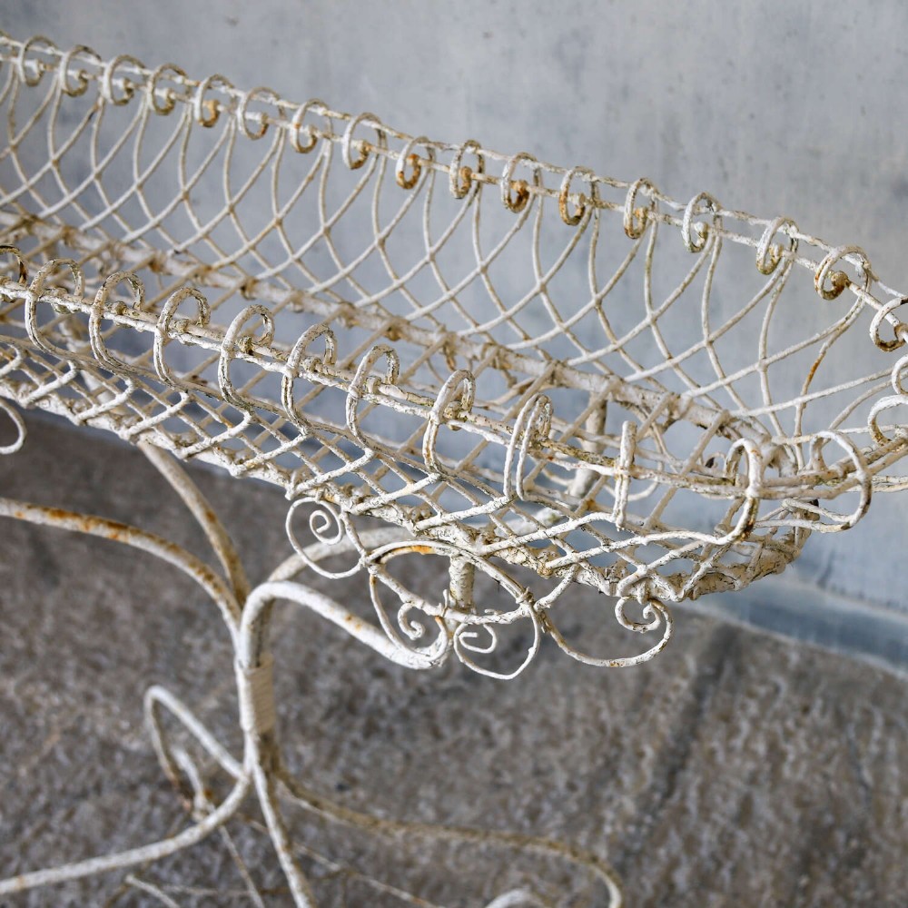 A 19th-century distressed white painted wrought iron plant stand featuring a wire lattice basket top over scrollwork legs. View V