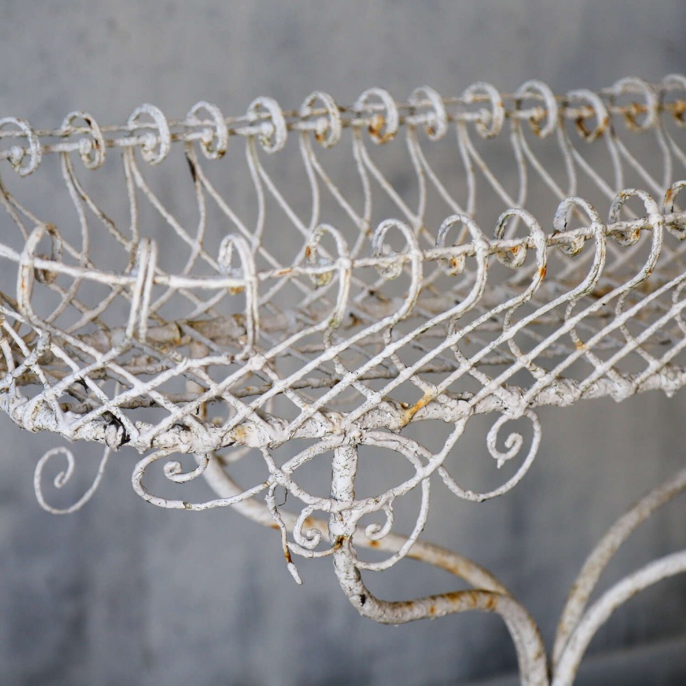 A 19th-century distressed white painted wrought iron plant stand featuring a wire lattice basket top over scrollwork legs. View VI