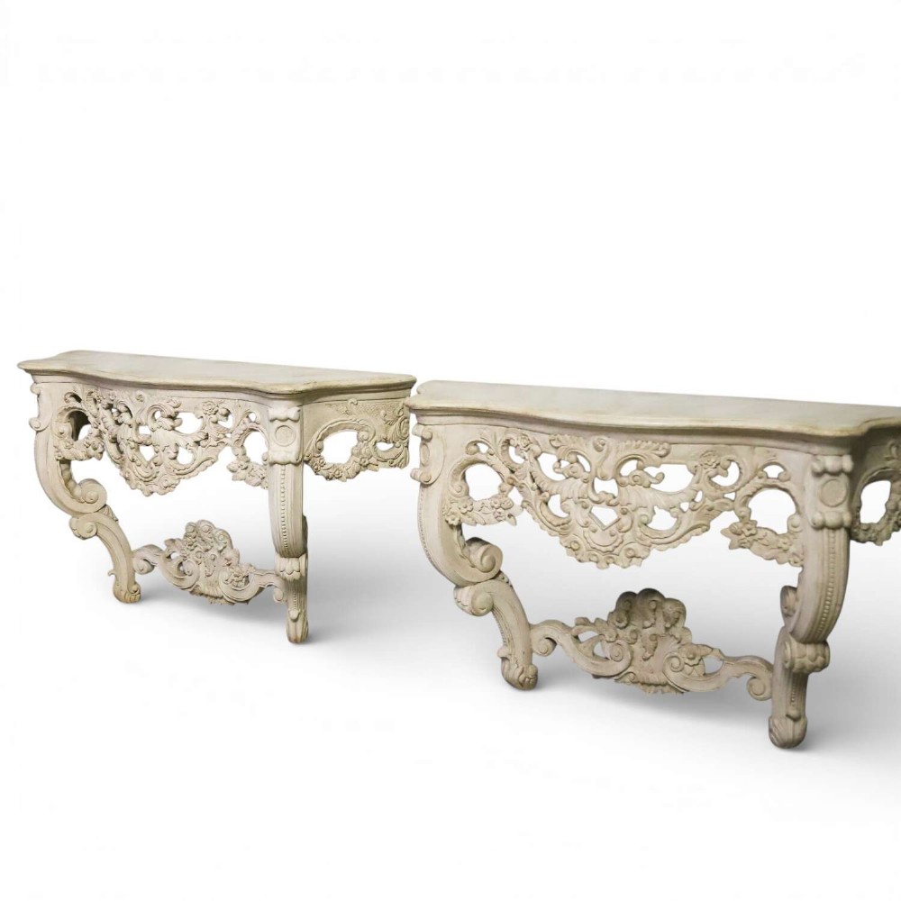 A pair of 1930s French wall console tables with intricate carved Rococo scrollwork in an antique white painted finish.