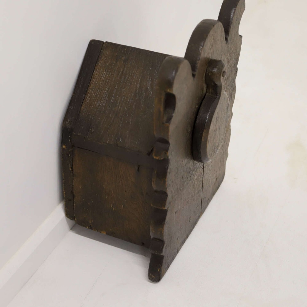 A dark brown 17th or 18th-century carved oak salt box featuring a shaped backboard with a circular hanging aperture and a sloping hinged lid. View V
