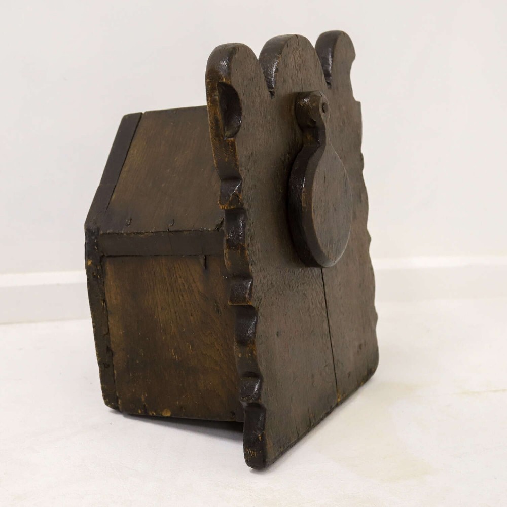 A dark brown 17th or 18th-century carved oak salt box featuring a shaped backboard with a circular hanging aperture and a sloping hinged lid. View XI