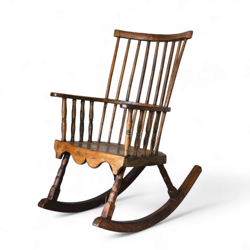 A 19th-century primitive comb-back rocking chair crafted from wood with tall back spindles, turned arm supports, and curved wooden runners.