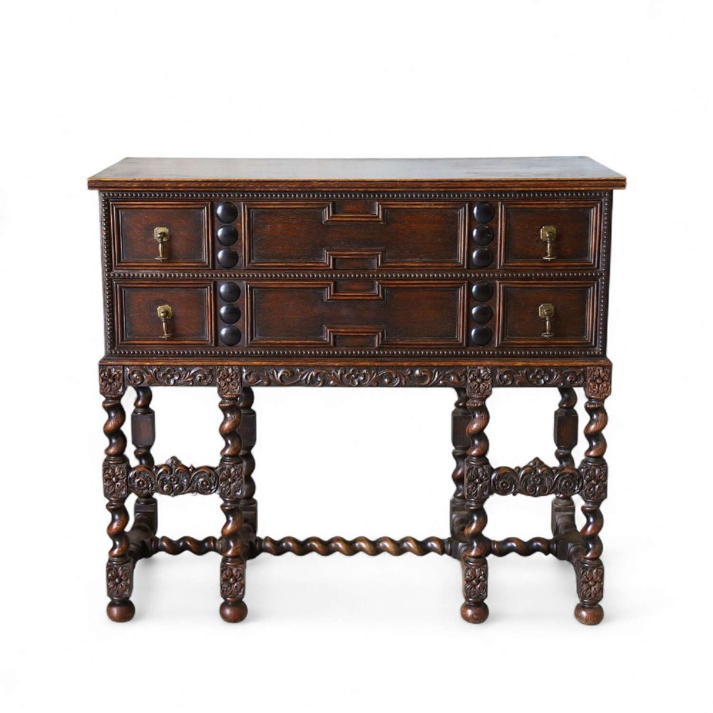 A dark oak Carolean Revival sideboard featuring two geometrically panelled drawers, intricate carving, and barley-twist legs connected by stretchers.