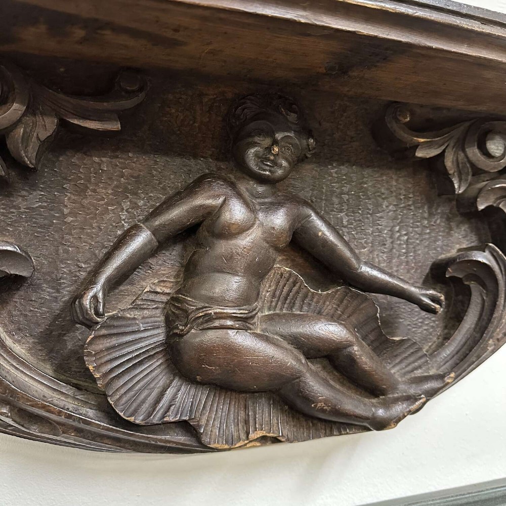 A dark oak wall shelf featuring detailed relief carvings of a central figure flanked by foliate scrollwork. View IV