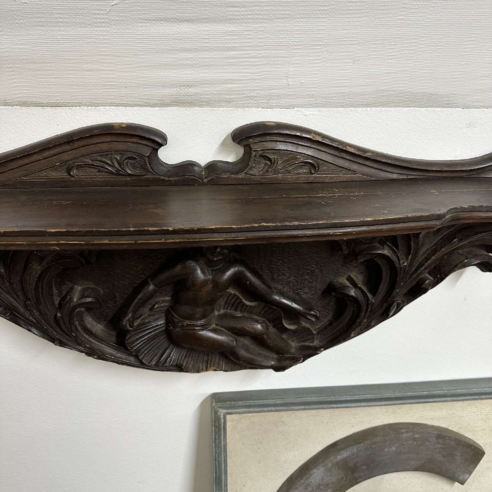 A dark oak wall shelf featuring detailed relief carvings of a central figure flanked by foliate scrollwork. View V