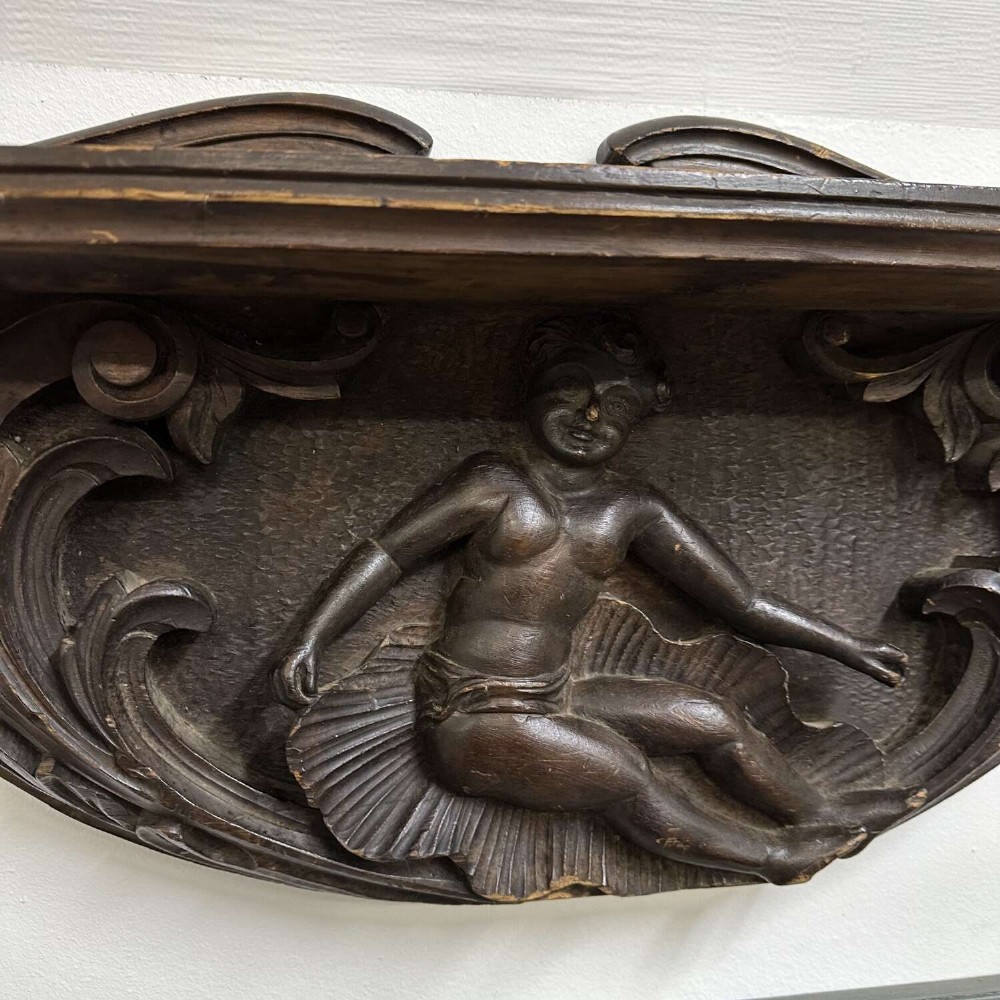 A dark oak wall shelf featuring detailed relief carvings of a central figure flanked by foliate scrollwork. View VI