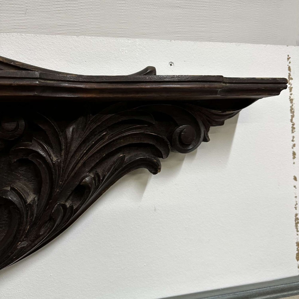 A dark oak wall shelf featuring detailed relief carvings of a central figure flanked by foliate scrollwork. View VII