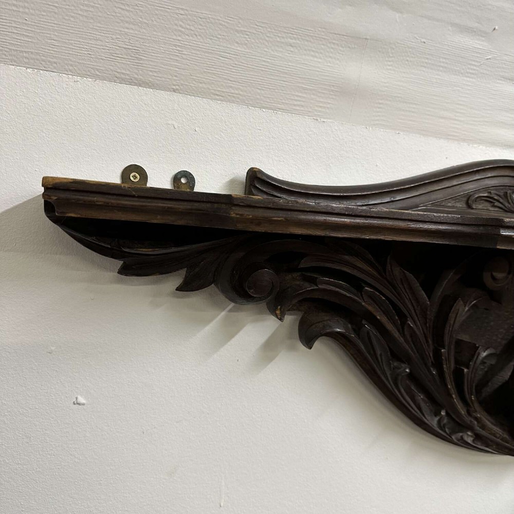 A dark oak wall shelf featuring detailed relief carvings of a central figure flanked by foliate scrollwork. View VIII