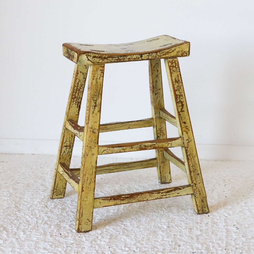 An antique Chinese elm stool featuring a slightly curved seat and a heavily distressed yellow lacquered finish over four splayed legs with horizontal stretchers.