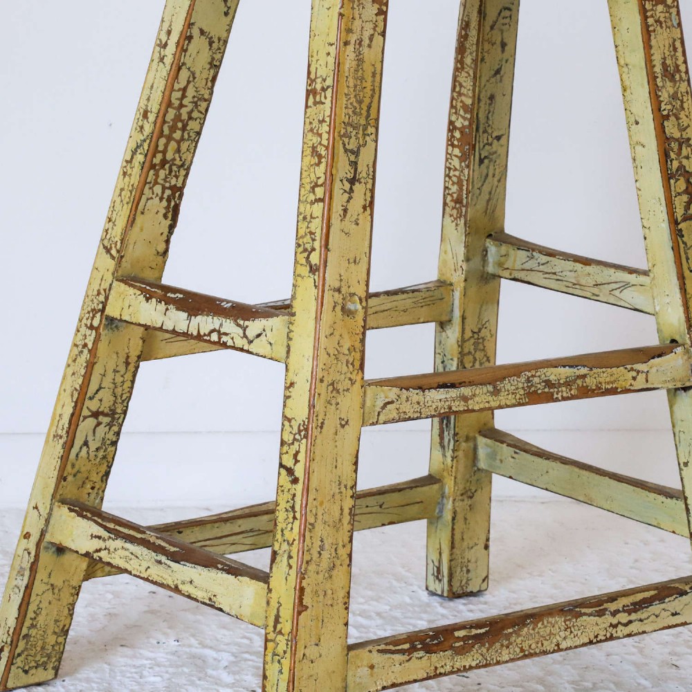 An antique Chinese elm stool featuring a slightly curved seat and a heavily distressed yellow lacquered finish over four splayed legs with horizontal stretchers. View V