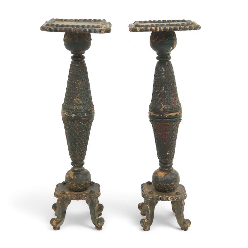 A pair of carved wooden torcheres featuring fish-scale carved baluster columns, square top platforms with beaded borders, scrolled feet, and distressed original green paint.