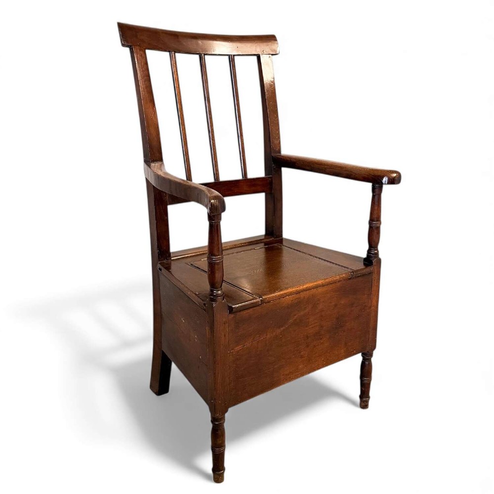 An early Georgian dark oak and walnut commode armchair featuring a vertical slatted back, curved armrests, turned front supports, and a deep enclosed box seat.