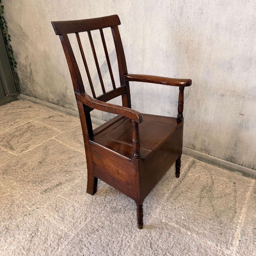 An early Georgian dark oak and walnut commode armchair featuring a vertical slatted back, curved armrests, turned front supports, and a deep enclosed box seat. View II