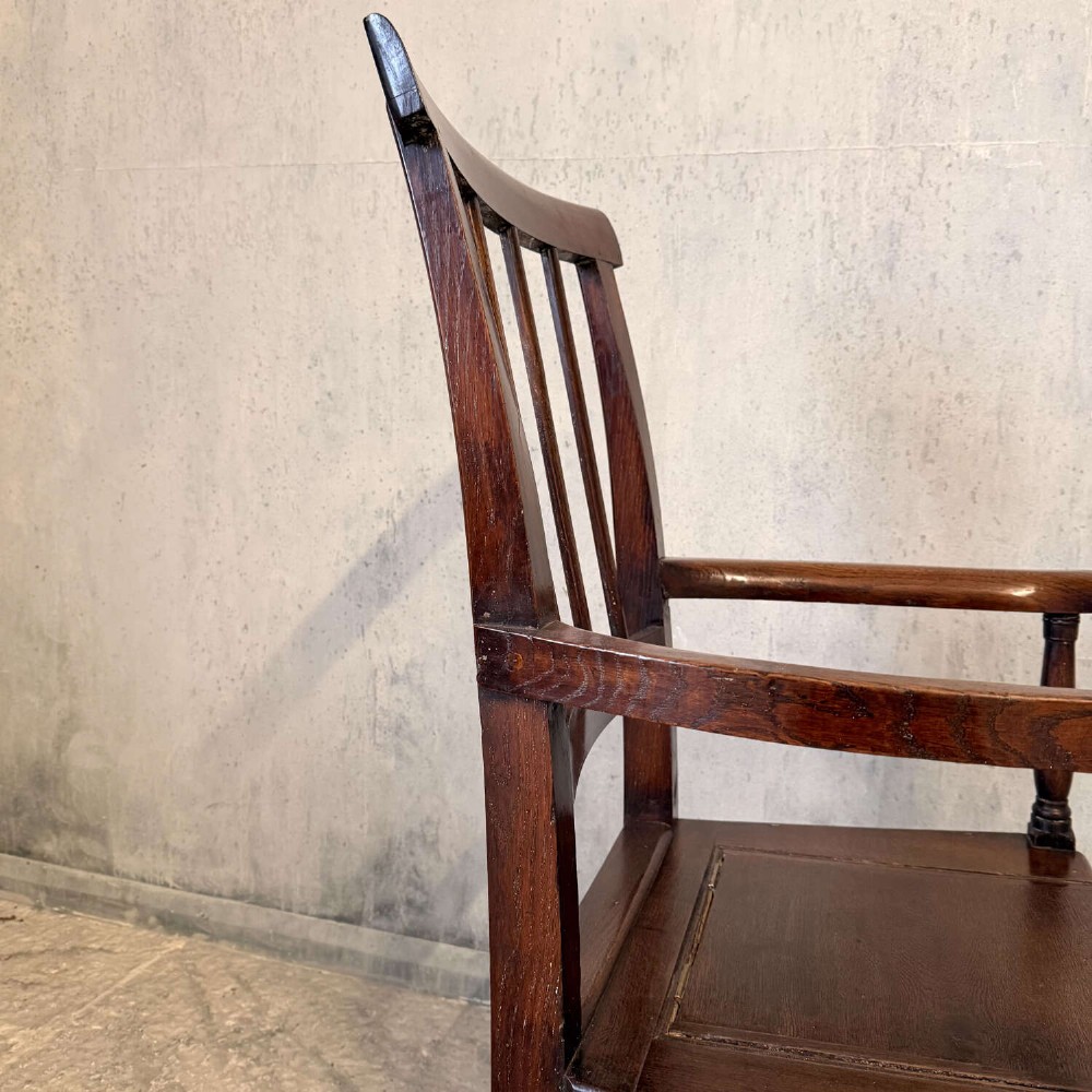 An early Georgian dark oak and walnut commode armchair featuring a vertical slatted back, curved armrests, turned front supports, and a deep enclosed box seat. View III