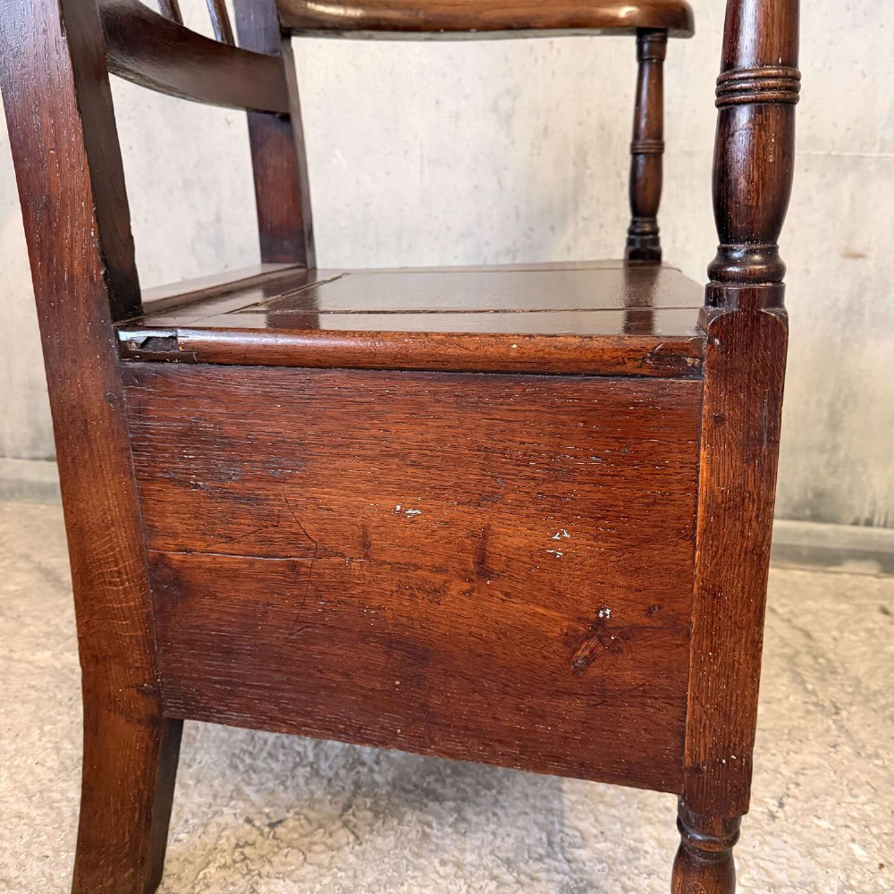 An early Georgian dark oak and walnut commode armchair featuring a vertical slatted back, curved armrests, turned front supports, and a deep enclosed box seat. View IV