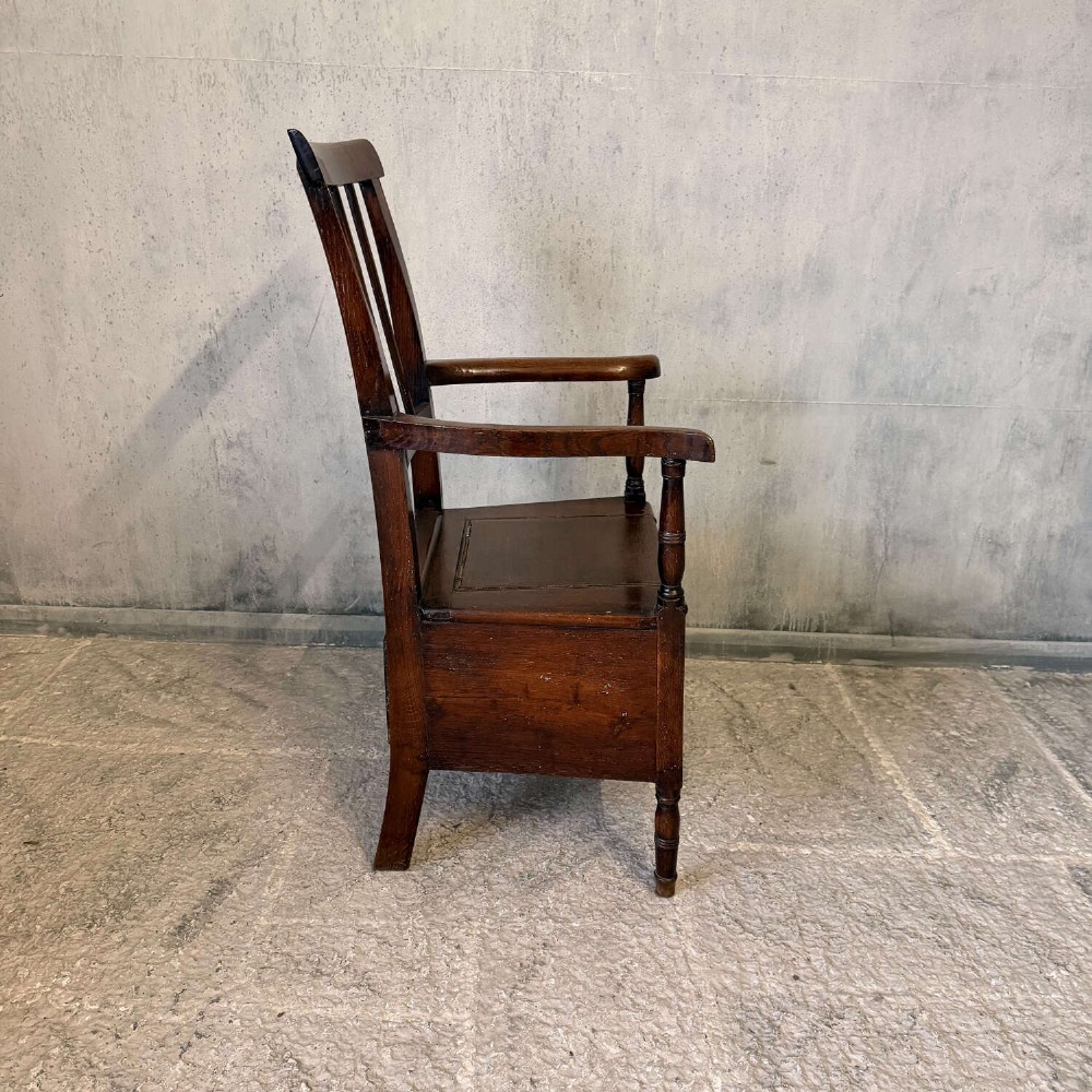 An early Georgian dark oak and walnut commode armchair featuring a vertical slatted back, curved armrests, turned front supports, and a deep enclosed box seat. View V
