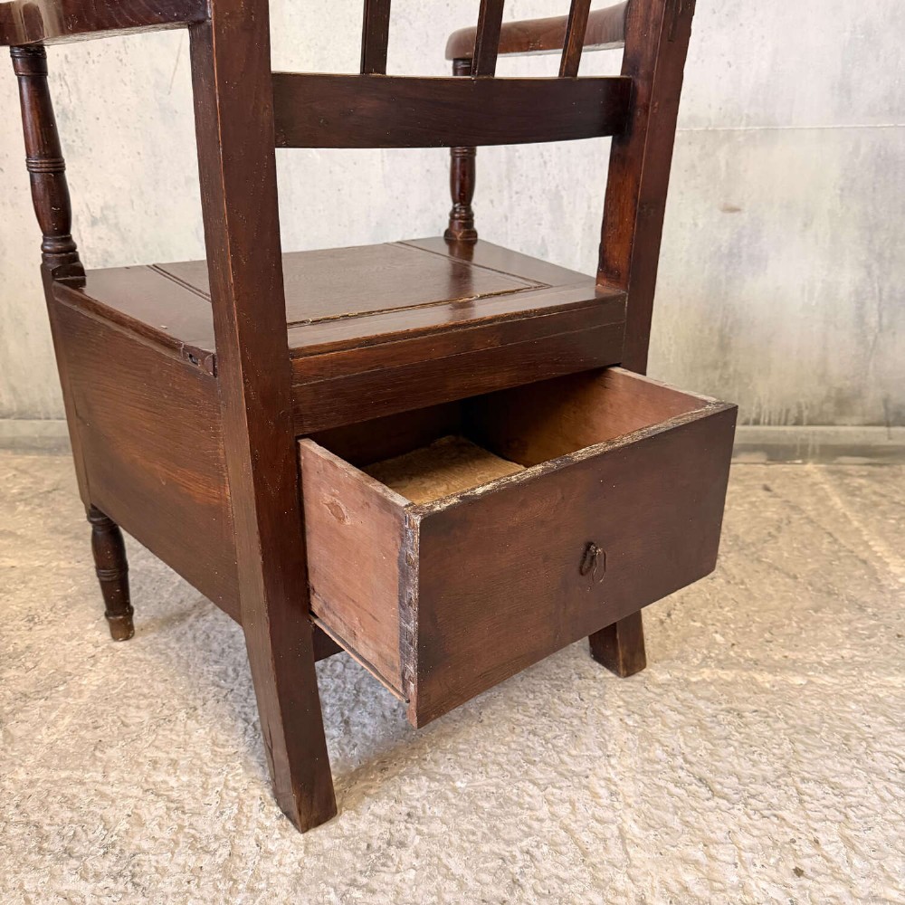 An early Georgian dark oak and walnut commode armchair featuring a vertical slatted back, curved armrests, turned front supports, and a deep enclosed box seat. View VI