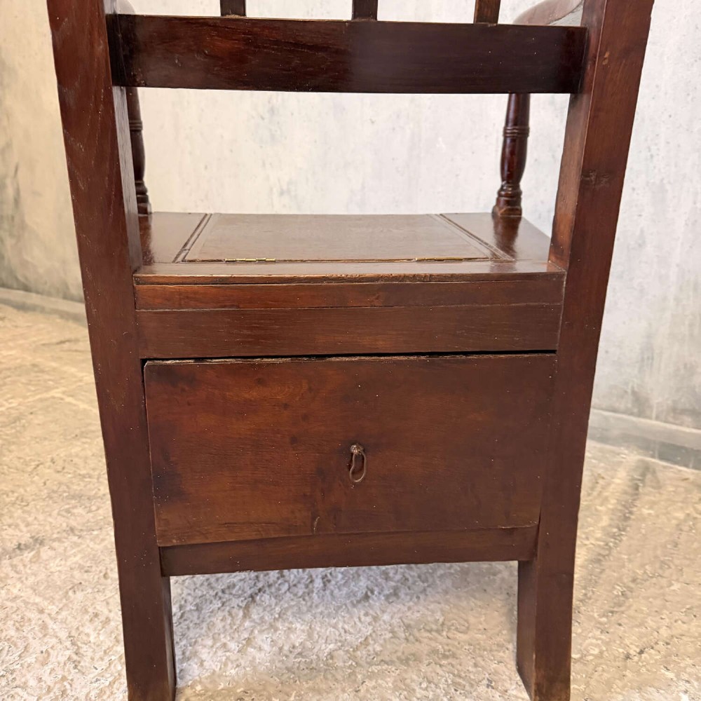 An early Georgian dark oak and walnut commode armchair featuring a vertical slatted back, curved armrests, turned front supports, and a deep enclosed box seat. View VII