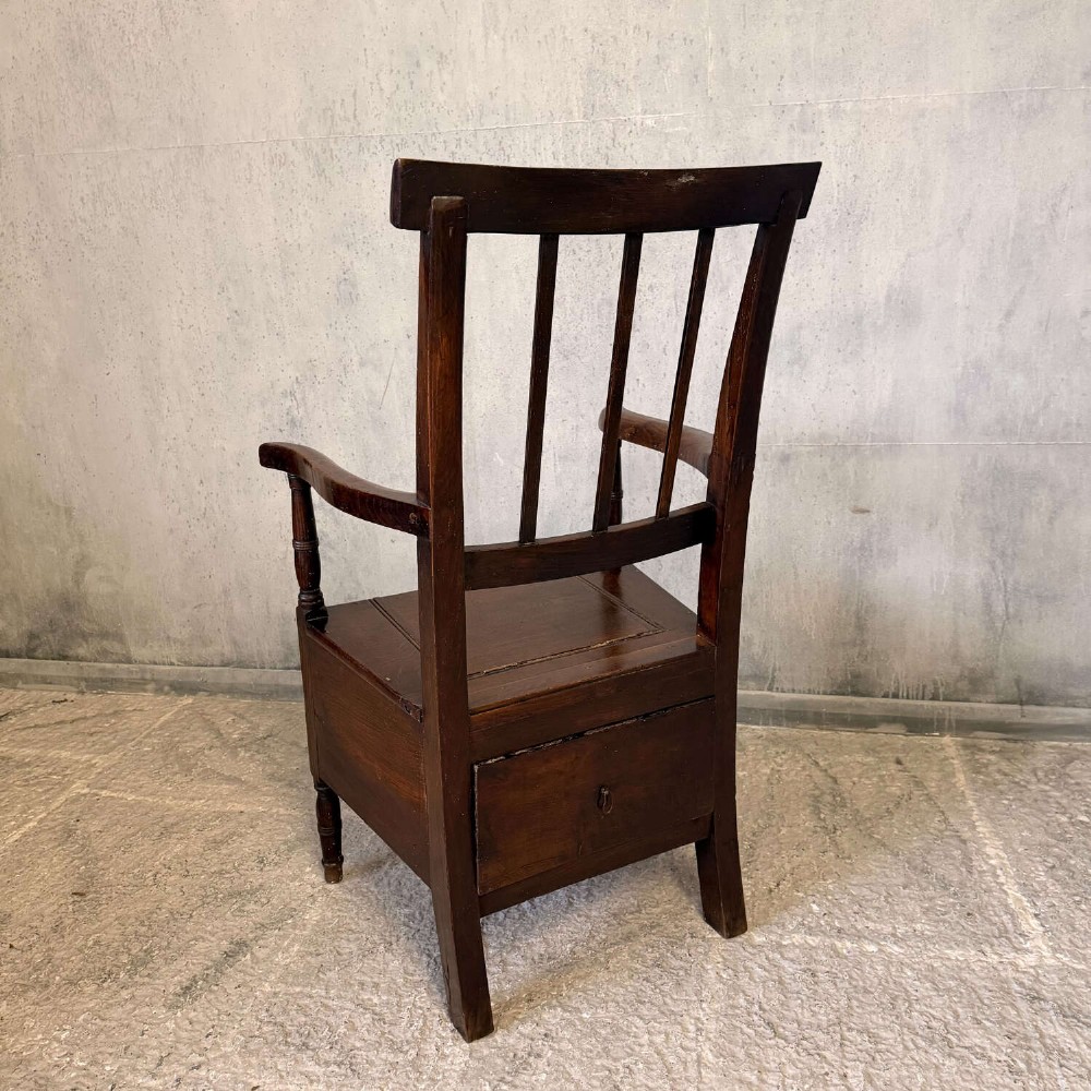 An early Georgian dark oak and walnut commode armchair featuring a vertical slatted back, curved armrests, turned front supports, and a deep enclosed box seat. View VIII