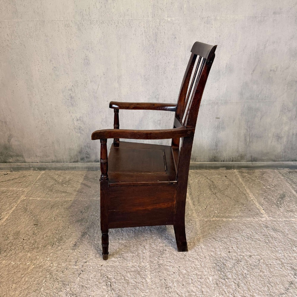 An early Georgian dark oak and walnut commode armchair featuring a vertical slatted back, curved armrests, turned front supports, and a deep enclosed box seat. View X
