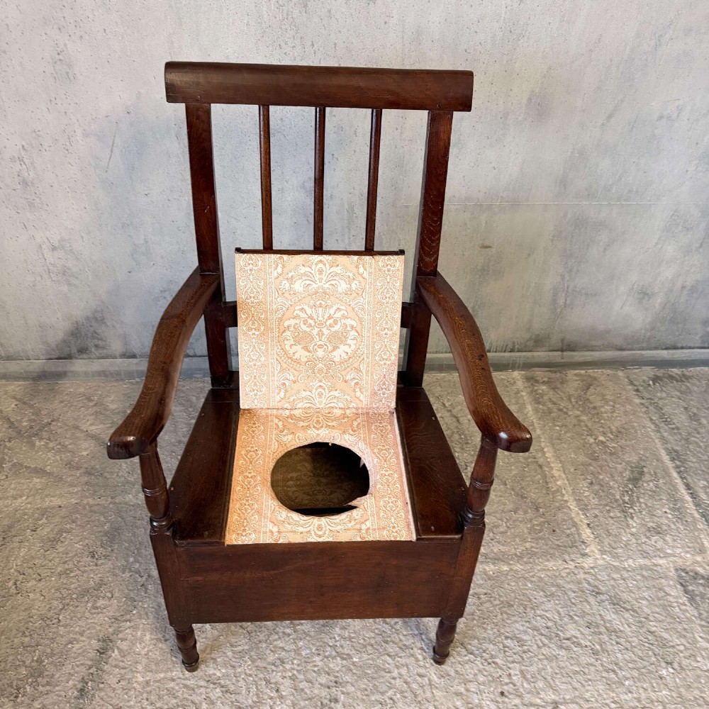 An early Georgian dark oak and walnut commode armchair featuring a vertical slatted back, curved armrests, turned front supports, and a deep enclosed box seat. View XI