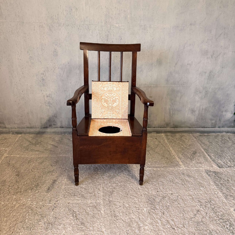 An early Georgian dark oak and walnut commode armchair featuring a vertical slatted back, curved armrests, turned front supports, and a deep enclosed box seat. View XII