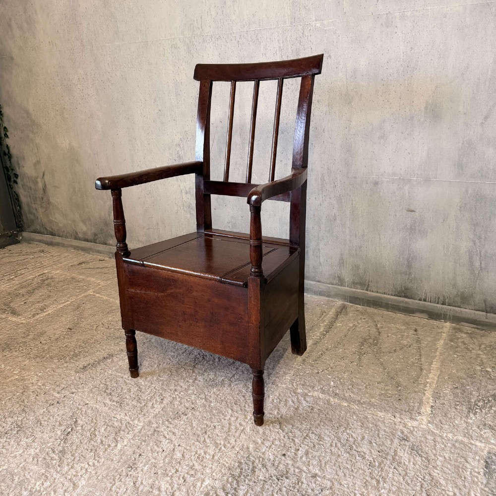 An early Georgian dark oak and walnut commode armchair featuring a vertical slatted back, curved armrests, turned front supports, and a deep enclosed box seat. View XIV