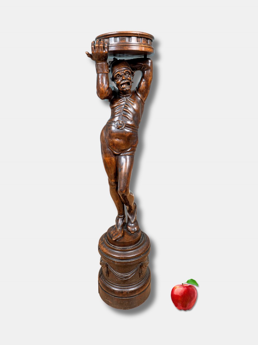 An Italian 19th-century hand-carved dark wood torchere stand depicting a jester figure holding a circular top on a moulded plinth base.