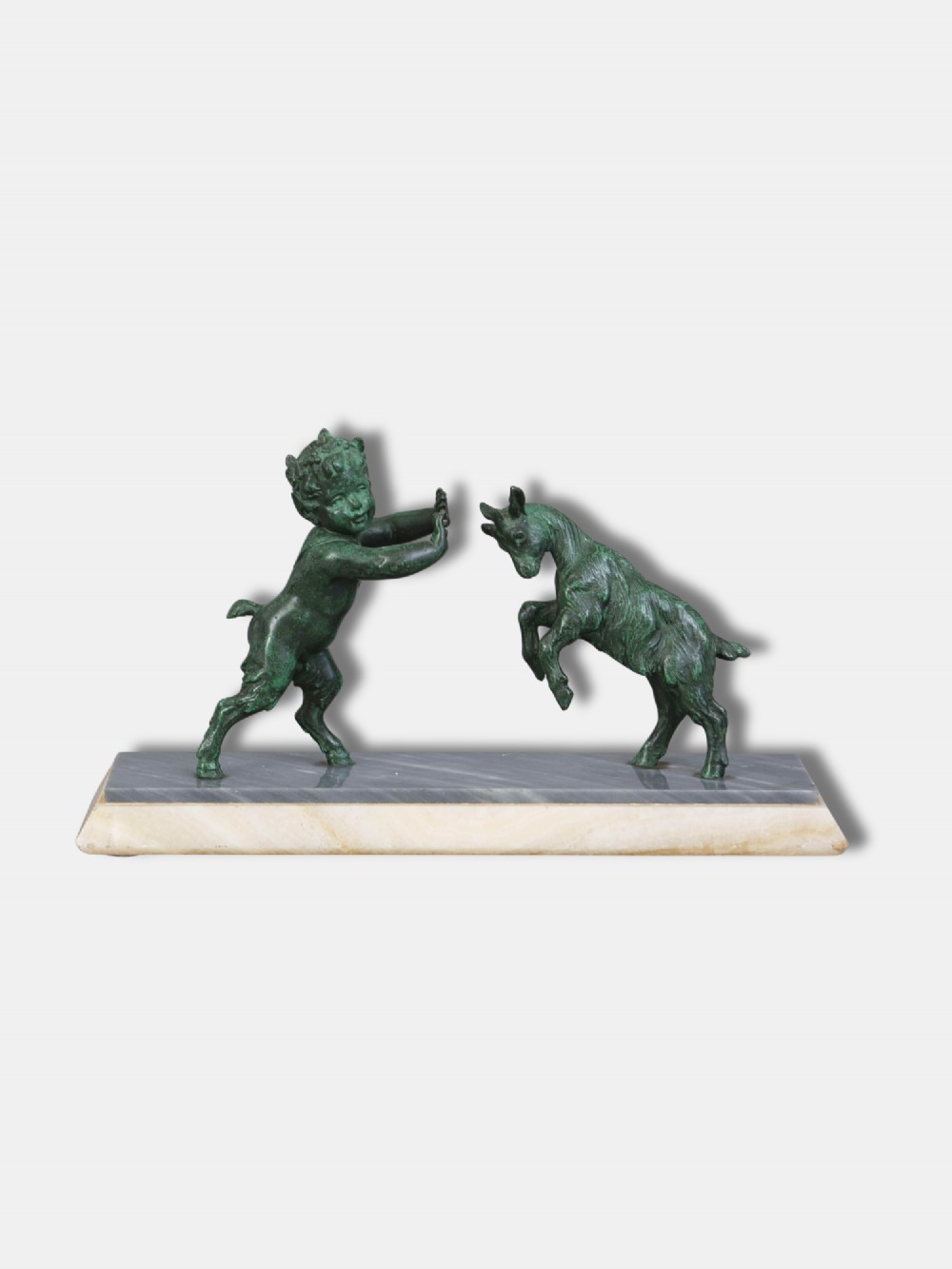 A green patinated bronze sculpture depicting a young faun interacting with a rearing goat, mounted on a rectangular marble base.