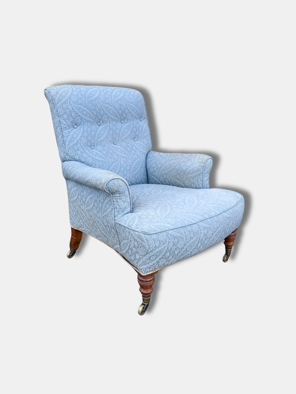 A late Victorian Howard and Sons armchair upholstered in light blue paisley fabric, featuring button-tufted backrest detail, rolled arms, and turned wooden front legs with brass castors.
