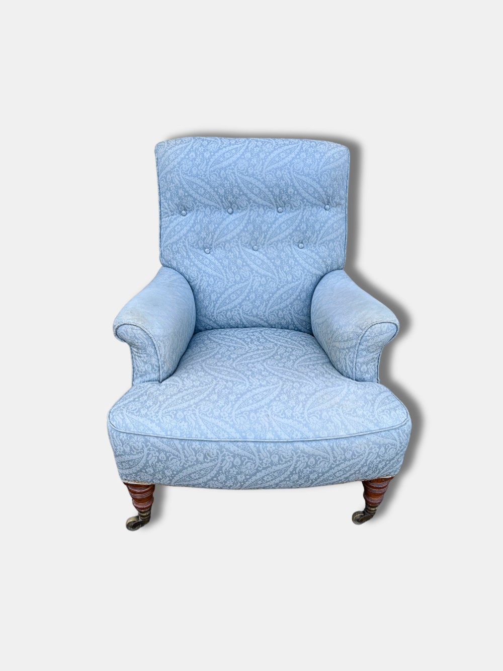 A late Victorian Howard and Sons armchair upholstered in light blue paisley fabric, featuring button-tufted backrest detail, rolled arms, and turned wooden front legs with brass castors. View II