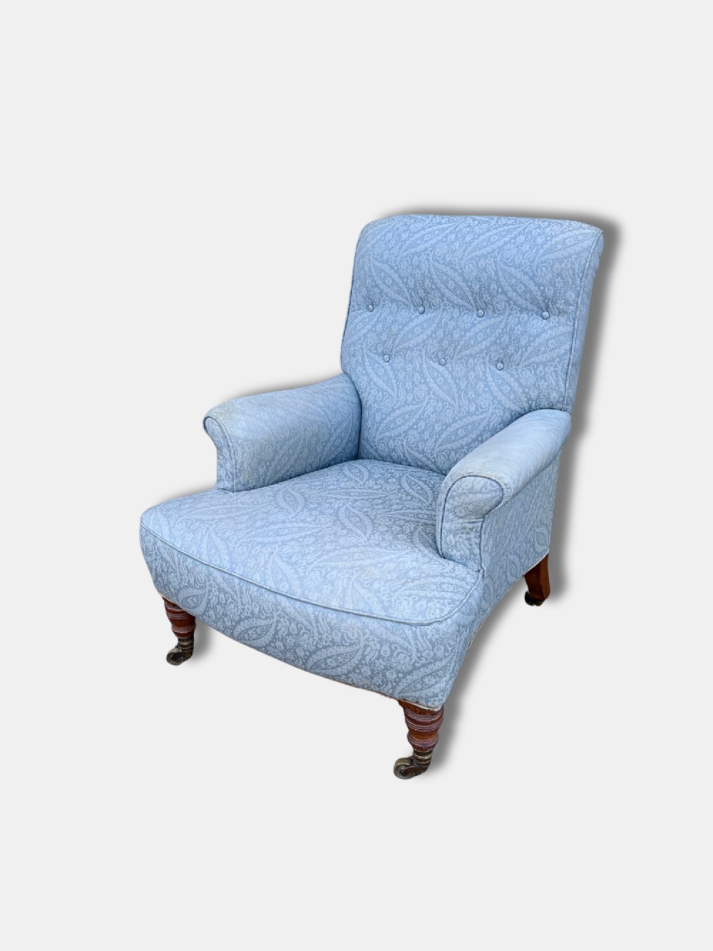 A late Victorian Howard and Sons armchair upholstered in light blue paisley fabric, featuring button-tufted backrest detail, rolled arms, and turned wooden front legs with brass castors. View III