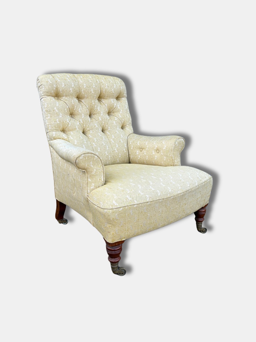 A Victorian armchair upholstered in pale yellow patterned fabric featuring a deep-buttoned backrest, scrolled armrests, and turned wood legs with brass castors.