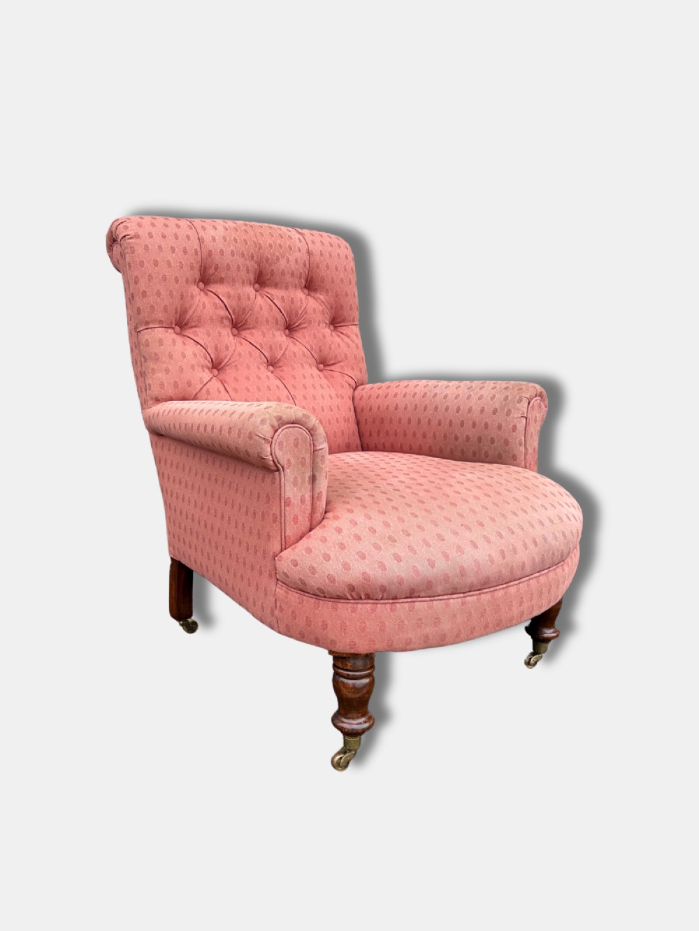 A late Victorian button-back armchair upholstered in dusty pink patterned fabric with low arms and turned wooden legs on brass castors.