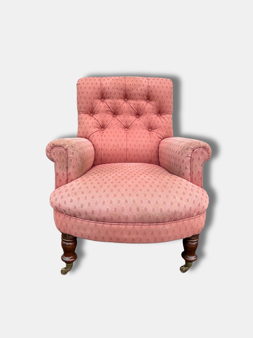 A late Victorian button-back armchair upholstered in dusty pink patterned fabric with low arms and turned wooden legs on brass castors. View II