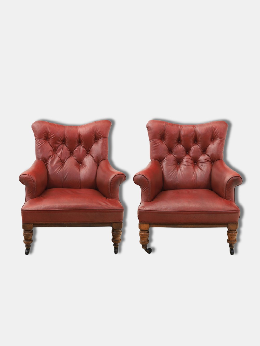 A pair of 1920s English armchairs upholstered in red oxblood rexine with deep buttoning and turned wooden legs on castors.