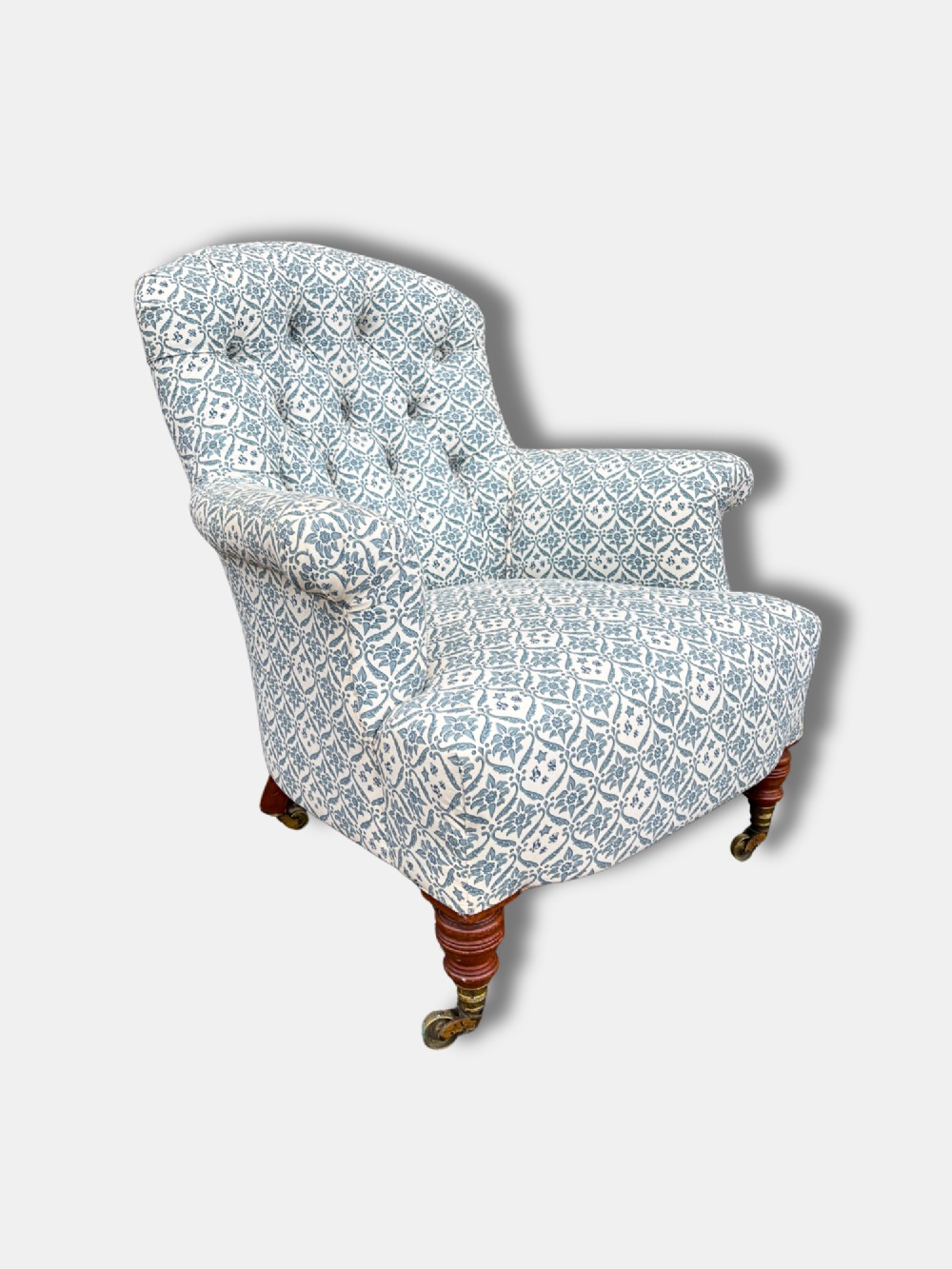 A late Victorian button back armchair upholstered in blue and white botanical patterned fabric, featuring turned wooden front legs on brass castors.