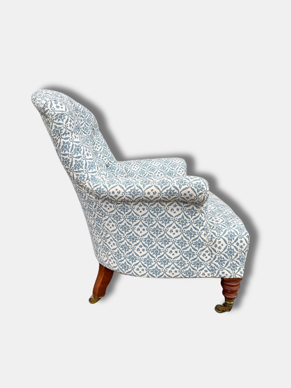 A late Victorian button back armchair upholstered in blue and white botanical patterned fabric, featuring turned wooden front legs on brass castors. View II