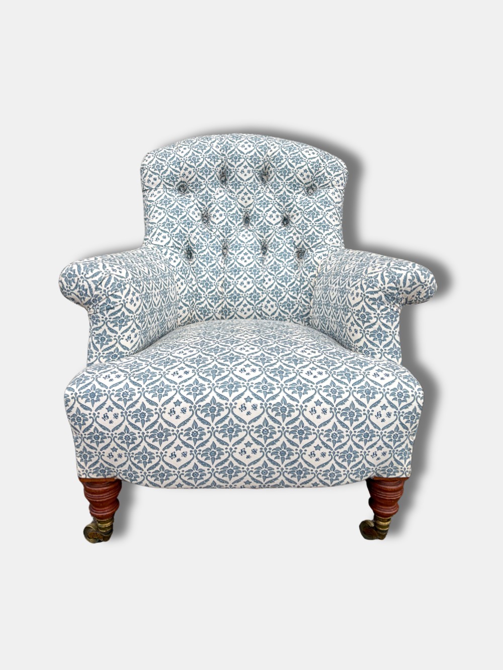 A late Victorian button back armchair upholstered in blue and white botanical patterned fabric, featuring turned wooden front legs on brass castors. View III