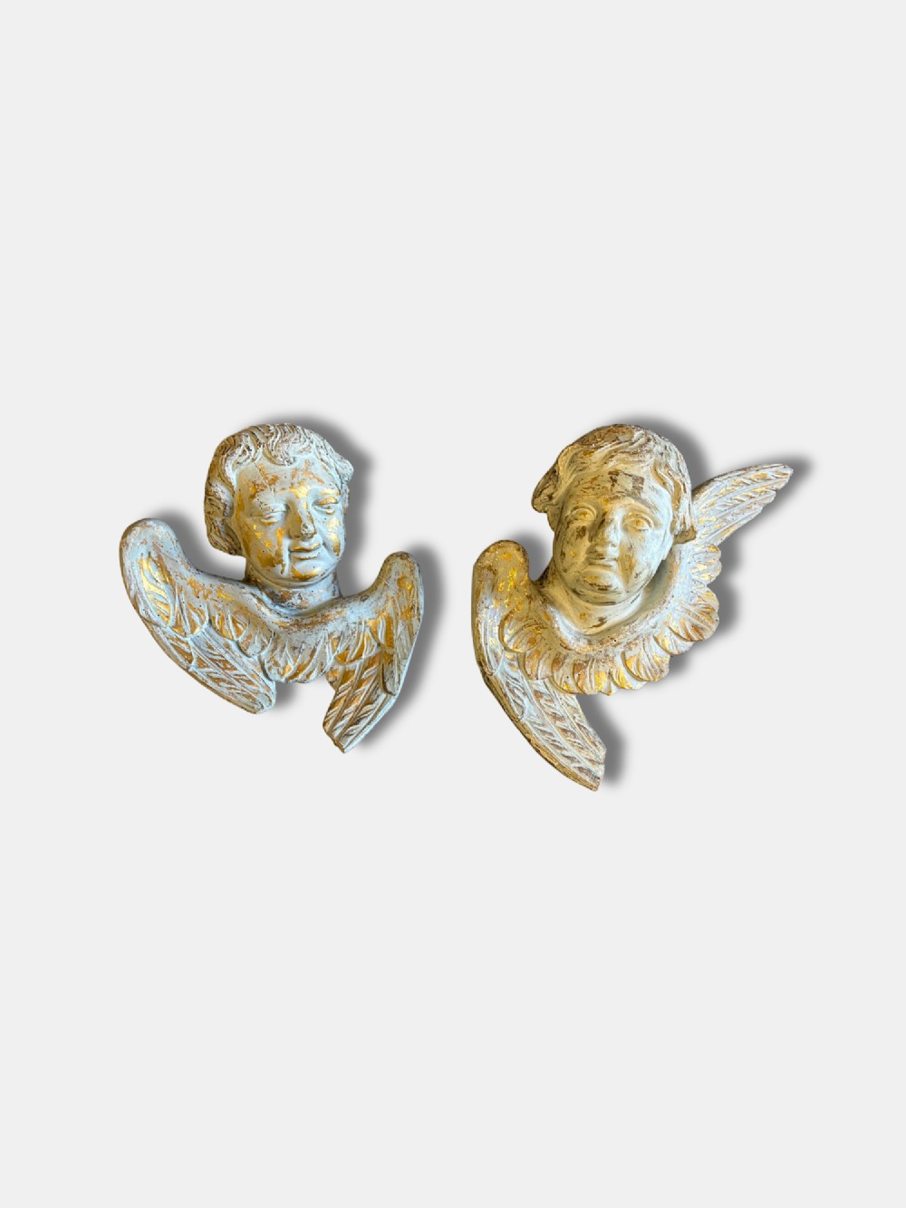 A pair of carved white and gilded French putti heads featuring winged detailing and distressed finishes.