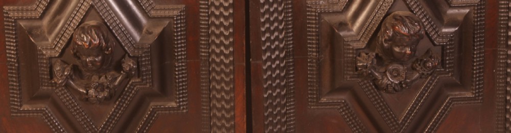 A pair of rectangular dark oak door panels featuring central raised diamond motifs carved with human heads and intricate geometric chip-carving borders. View III